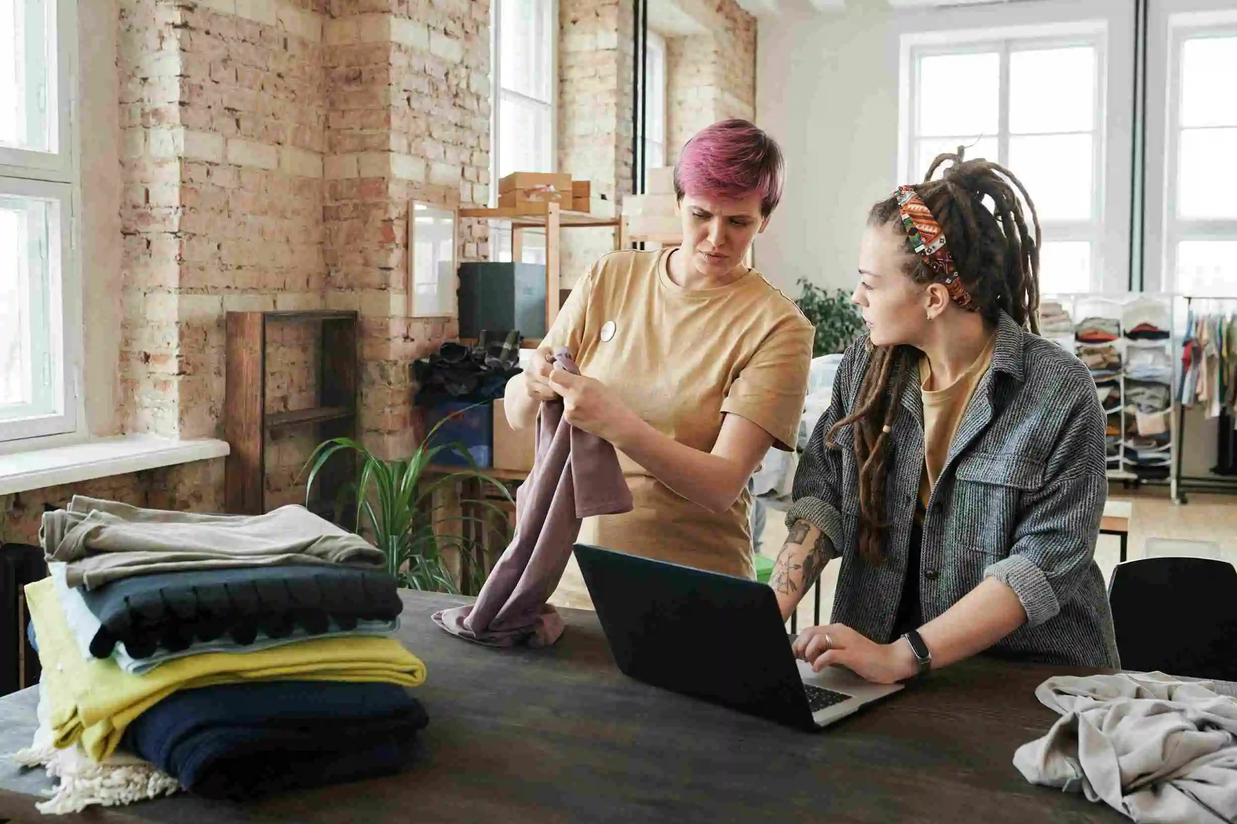 Two people are engaged in a conversation. One is showing the other something on a piece of clothing. The second person is typing on a laptop. A pile of folded cloth items sits on the table nearby.