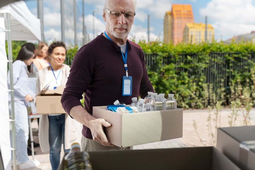 Male volunteer wearing a blue badge carries a box of bottled water and food supplies at an outdoor food bank distribution event,