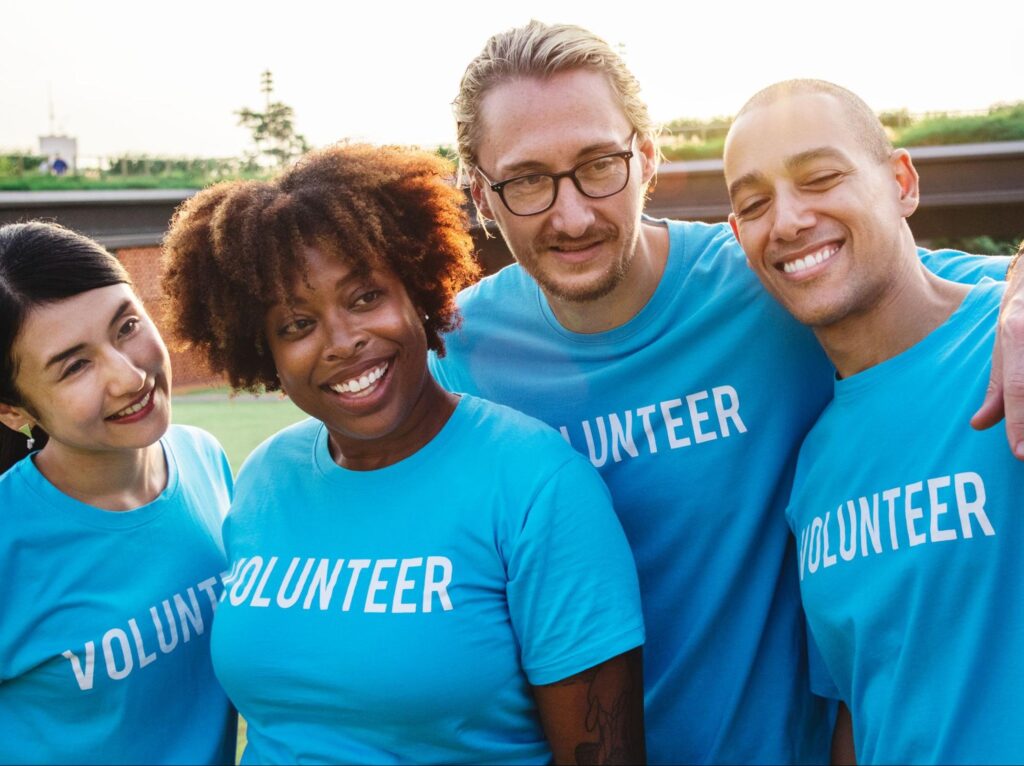 A diverse group of four smiling volunteers wearing matching blue shirts that say “VOLUNTEER,”
