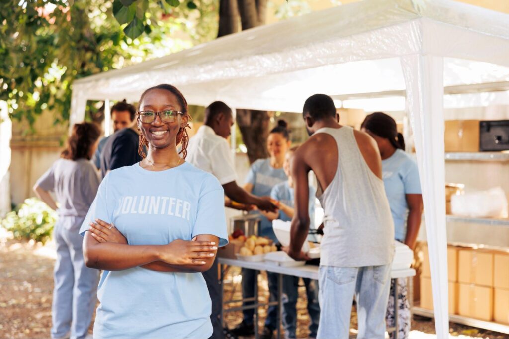 Young Black woman wearing glasses and a volunteer shirt smiling with arms crossed, as a team of volunteers serves food in the background.
