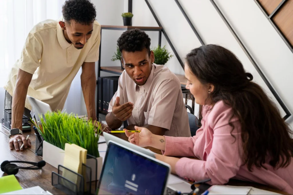 Three coworkers gathered around a desk in a bright office, collaborating on a project and reviewing notes together on a laptop.