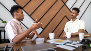 Two men sitting at a modern wooden desk having a focused discussion over coffee, with notebooks, a laptop, and office supplies on the table.
