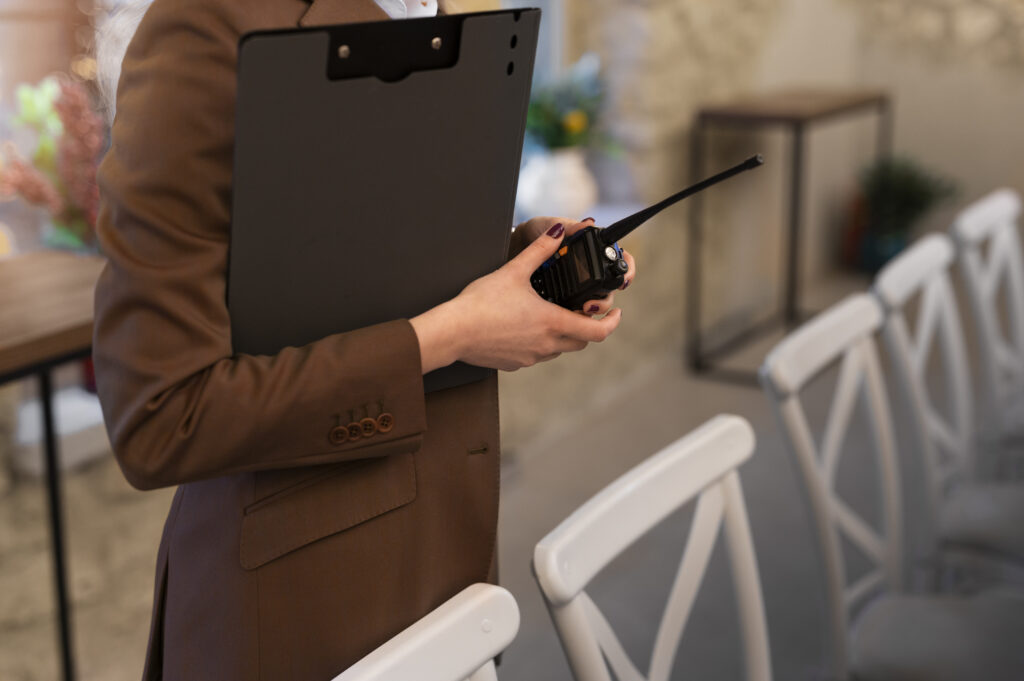 An individual standing behind a dining setup with a clipboard and walkie-talkie.